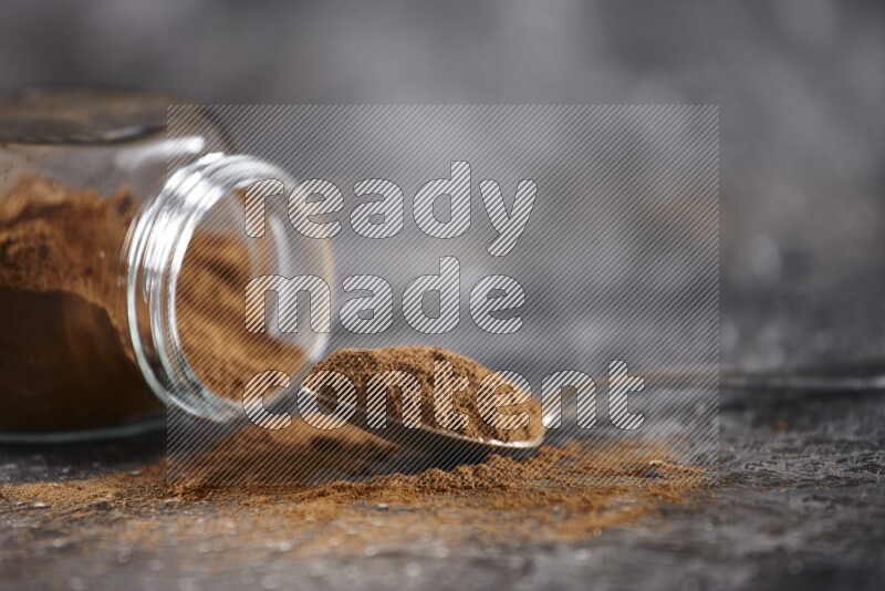 Herbal glass jar full of cinnamon powder flipped and a metal spoon on textured black background