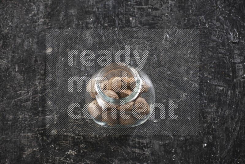 A glass spice jar full of whole nutmeg seeds on a textured black flooring