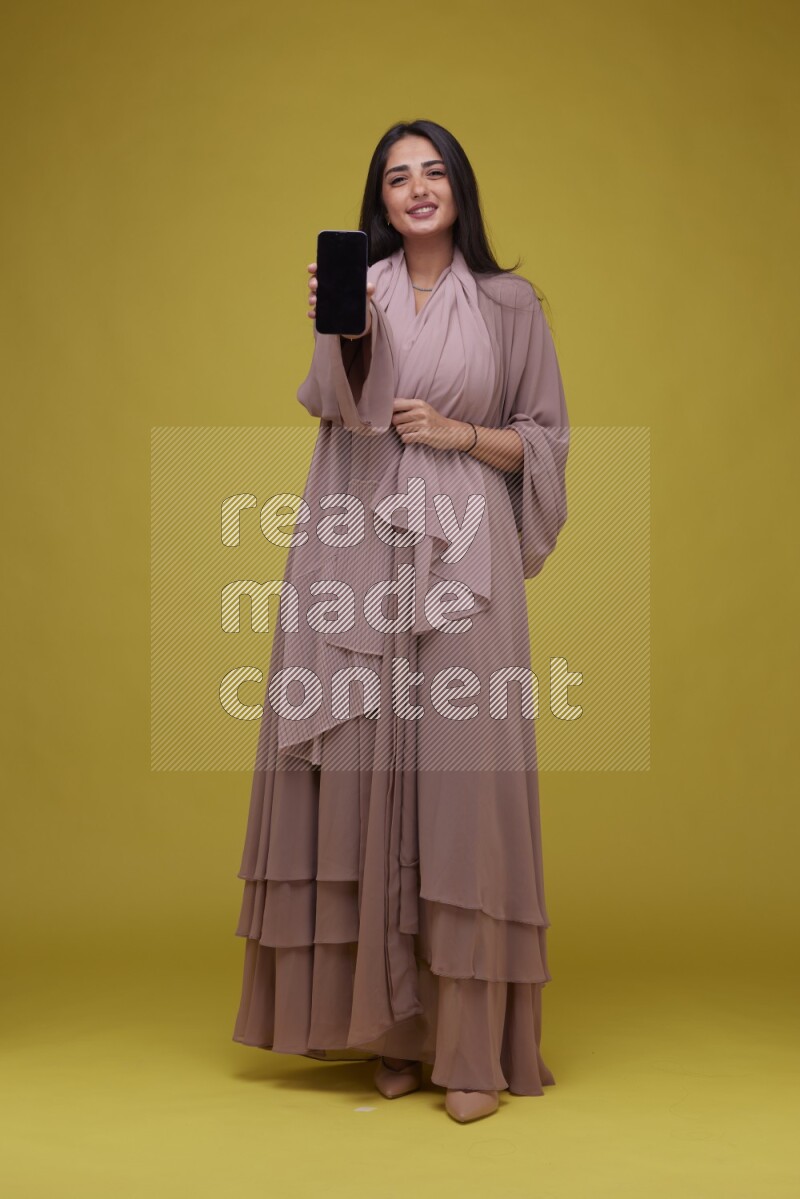 A woman Showing SmartPhone Screen on a Yellow Background wearing Brown Abaya