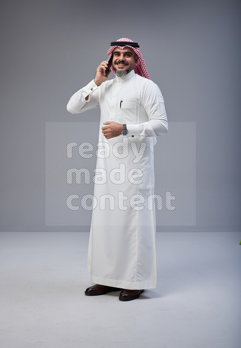 Saudi man Wearing Thob and red Shomag standing talking on phone on Gray background