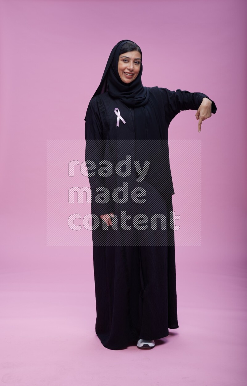 Saudi woman wearing pink ribbon on Abaya standing interacting with the camera on pink background
