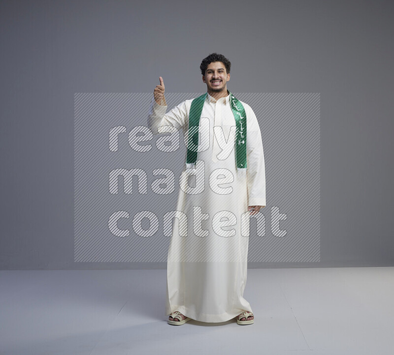 A Saudi man standing wearing thob and Saudi flag scarf on gray background