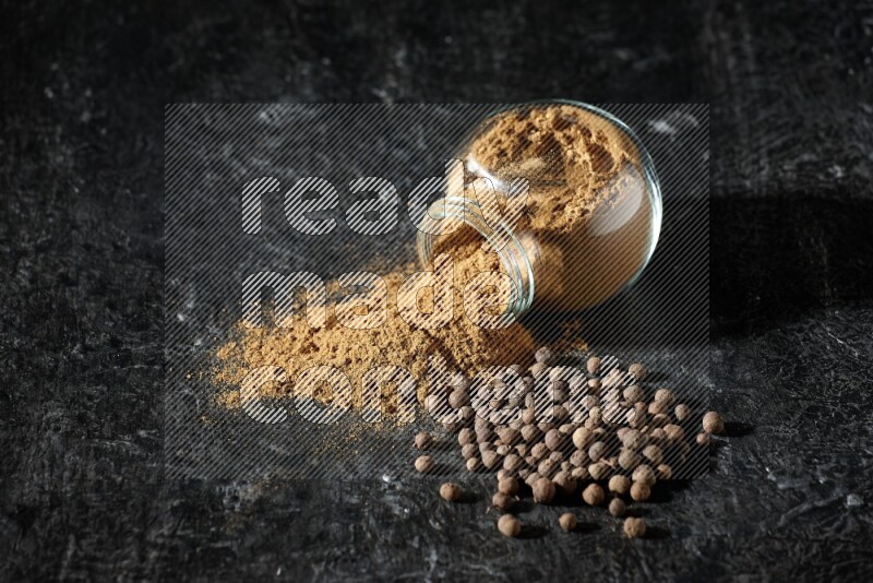 وعاء زجاجي للتوابل مقلوب ممتلئ ببودرة بهار الفلفل الحلو والبودرة تناثرت منه مع الحبوب على خلفية سوداء