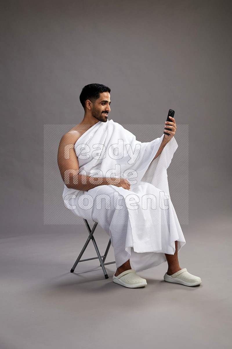 A man wearing Ehram sitting on chair taking selfie on gray background