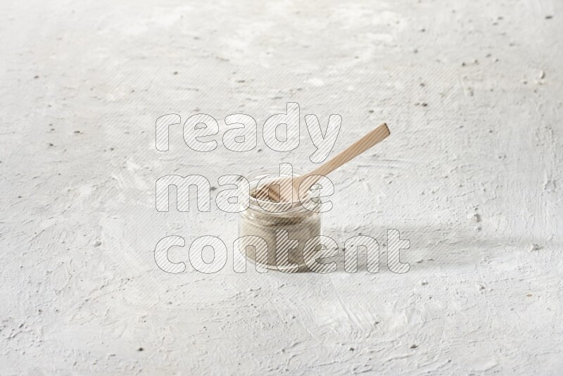 A glass jar and wooden spoon full of white pepper powder on textured white flooring