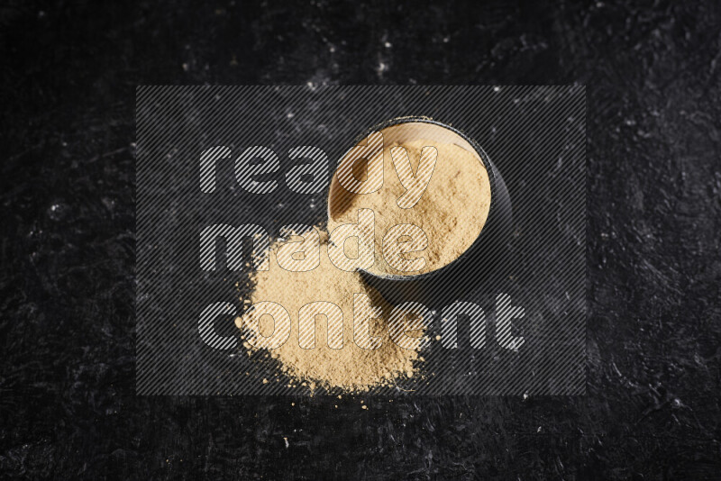 A black pottery bowl full of ground ginger powder with fallen powder from it on black background