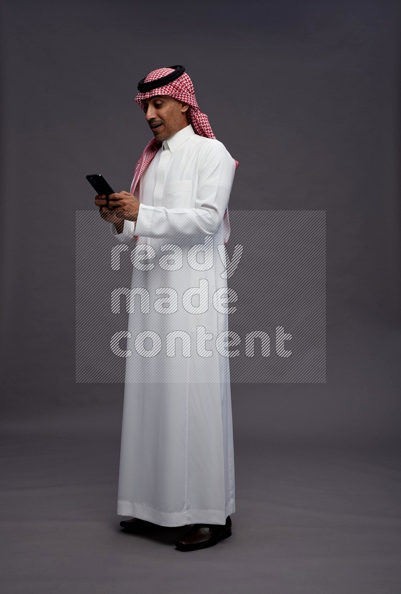 Saudi man wearing thob and shomag standing texting on phone on gray background