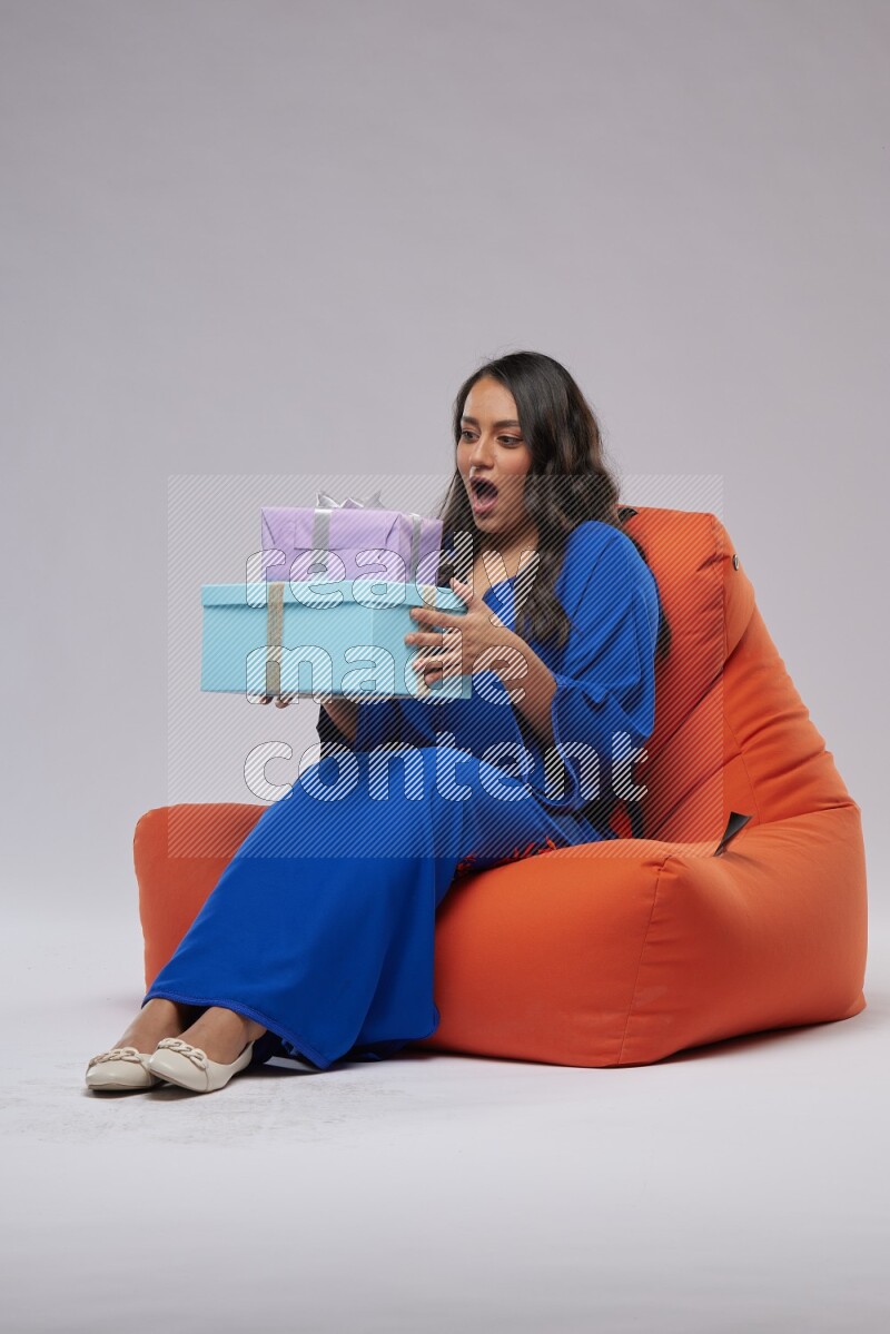 A woman sitting on an orange beanbag wearing Jalabeya holding a gift box