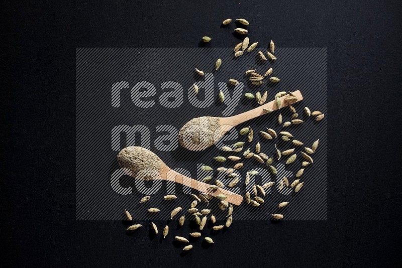 2 Wooden spoons full of cardamom powder and cardamom seeds spreaded on black flooring