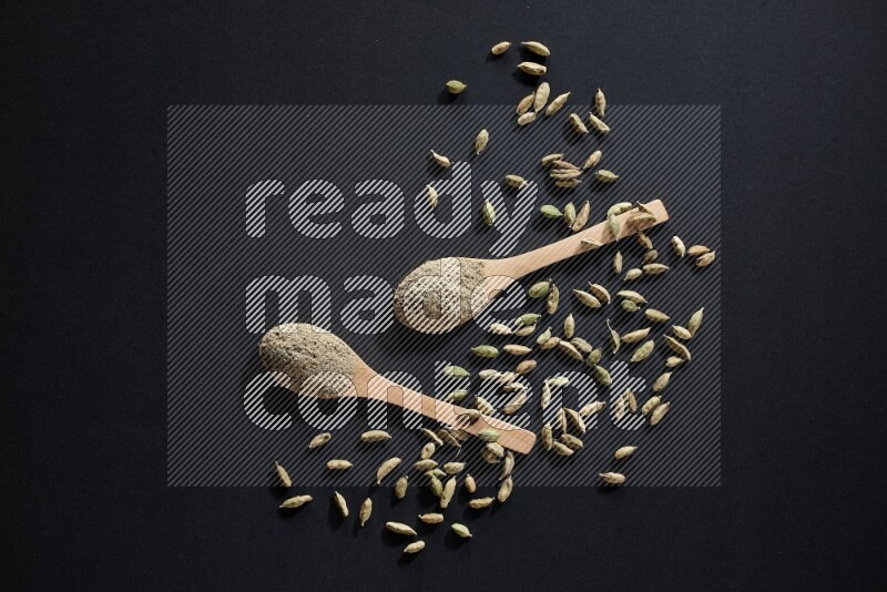 2 Wooden spoons full of cardamom powder and cardamom seeds spreaded on black flooring