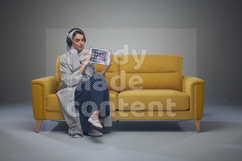 A Saudi female wearing a light gray Abaya and head scarf sitting on a yellow sofa and showing her tablet's screen while wearing headphones eye level on a grey background