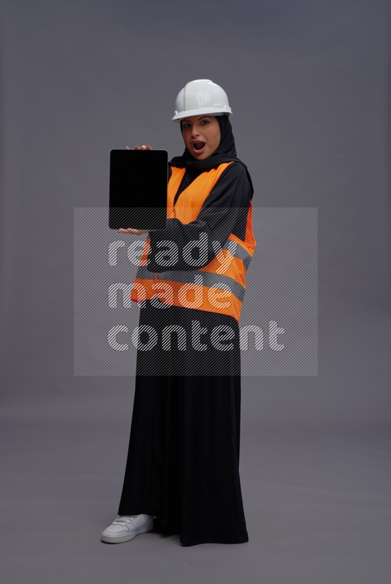 Saudi woman wearing Abaya with engineer vest standing showing tablet to camera on gray background