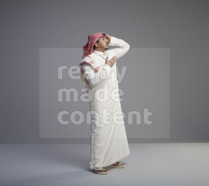 A Saudi man standing wearing thob and red shomag interacting with the camera on gray background