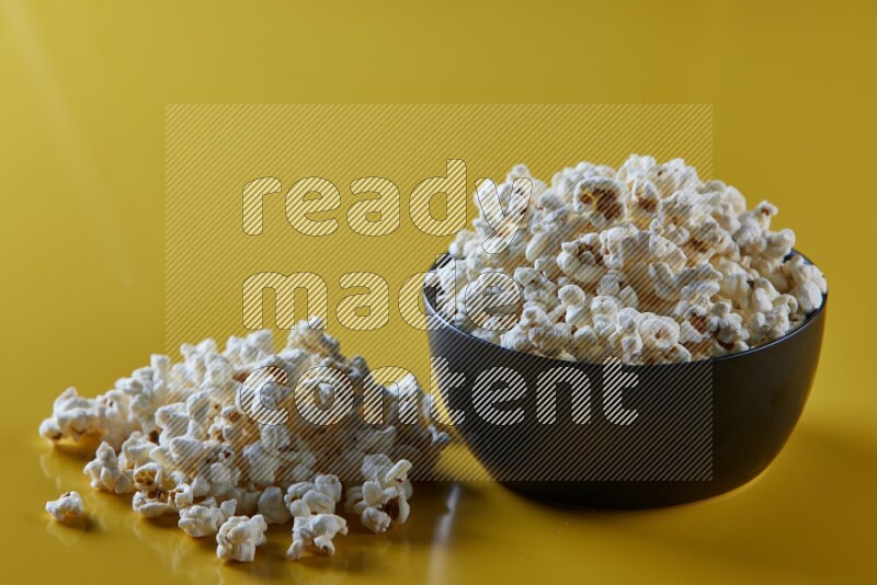 A black ceramic bowl full of popcorn with popcorn beside it on a yellow background in different angles