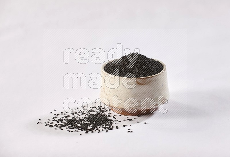 A beige pottery bowl full of black seeds and more seeds spread on a white flooring