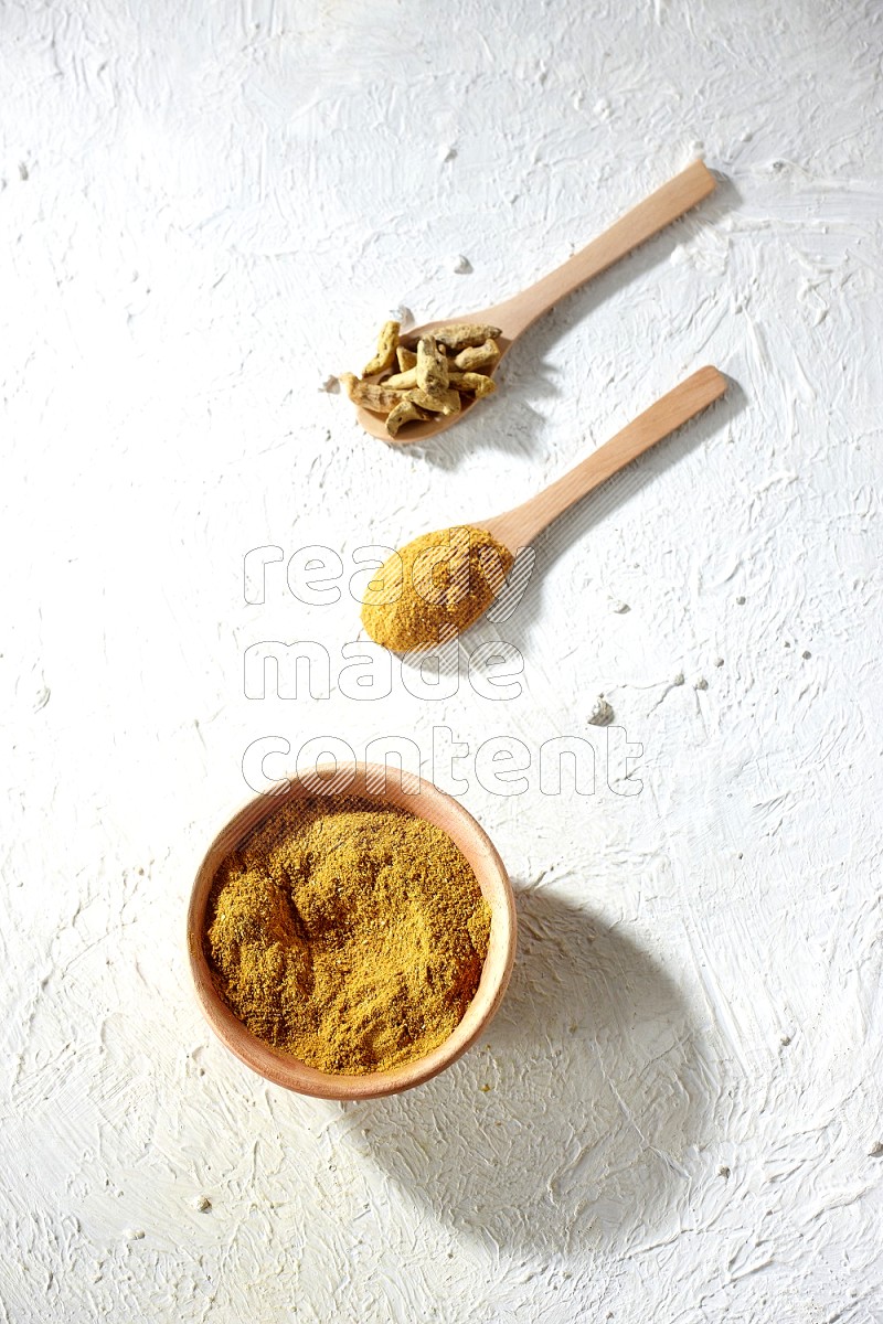 A wooden bowl full of turmeric powder and 2 wooden spoons full of dried turmeric whole finger and turmeric powder on textured white flooring