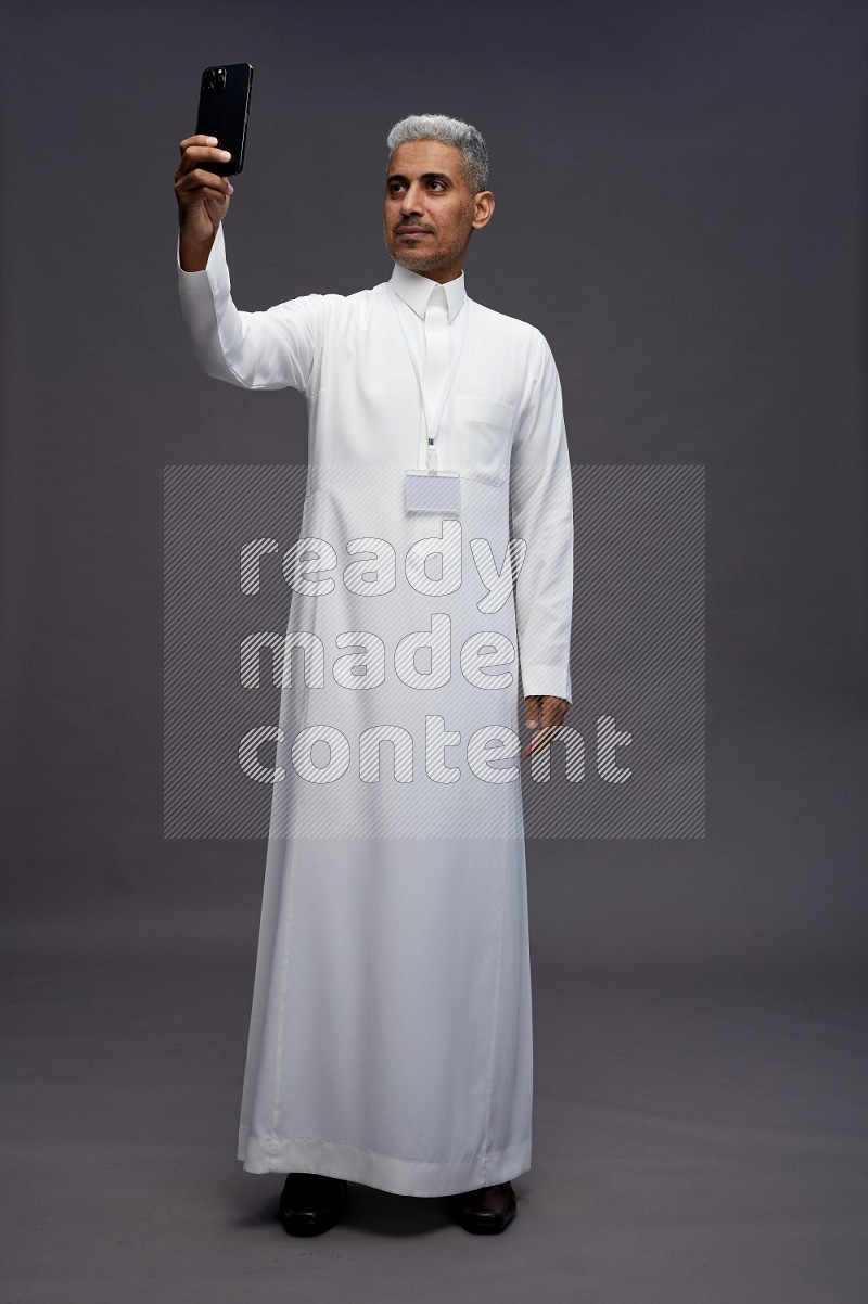 Saudi man wearing thob with neck strap employee badge standing taking selife on gray background