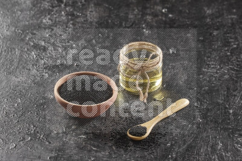 A wooden bowl and spoon full of black seeds and a glass jar of black seeds oil on a textured black flooring