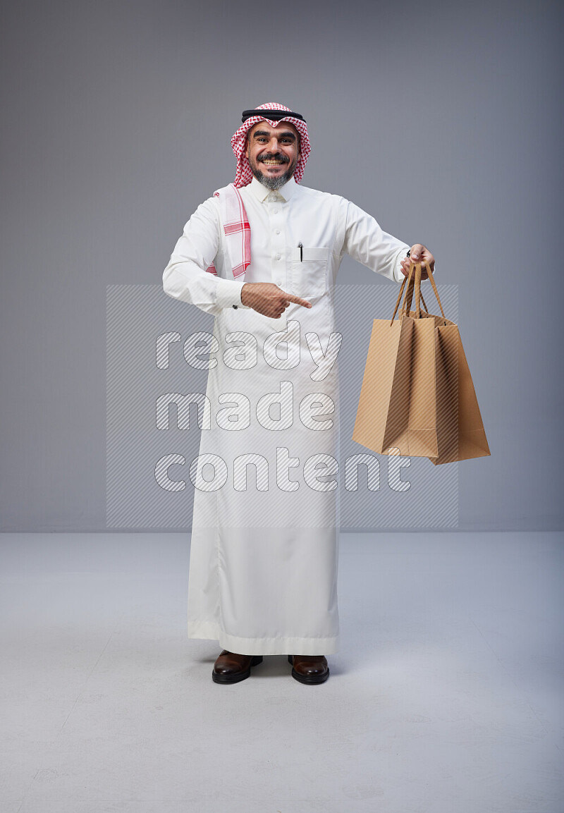 Saudi man Wearing Thob and red Shomag standing holding shopping bag on Gray background