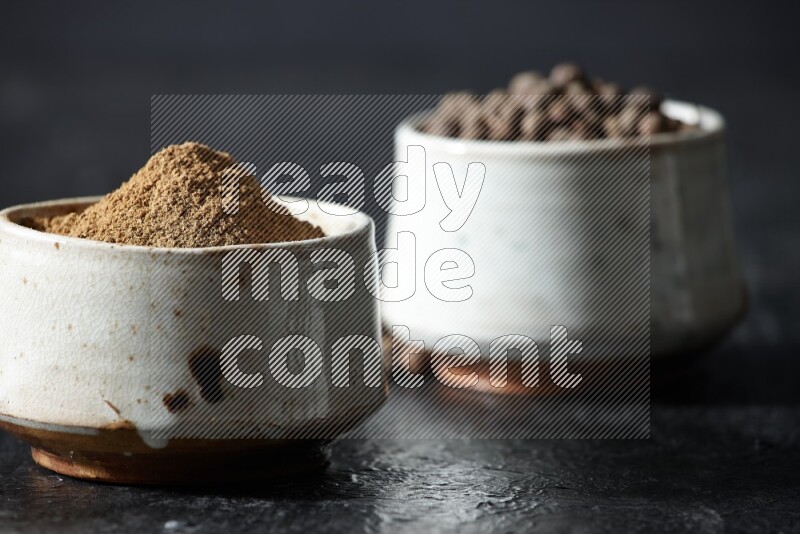 2 beige bowls, one full of allspice powder and the other full of whole balls on a textured black flooring