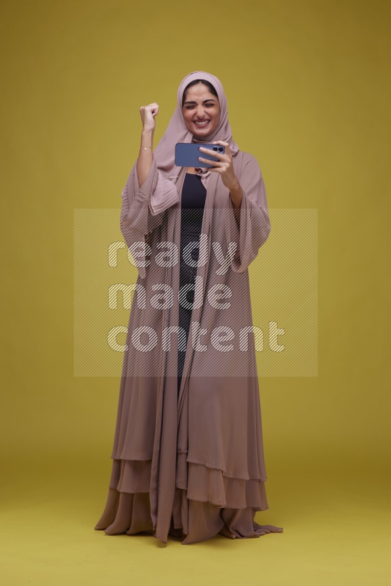 A woman Playing Games a Yellow Background wearing Brown Abaya with Hijab
