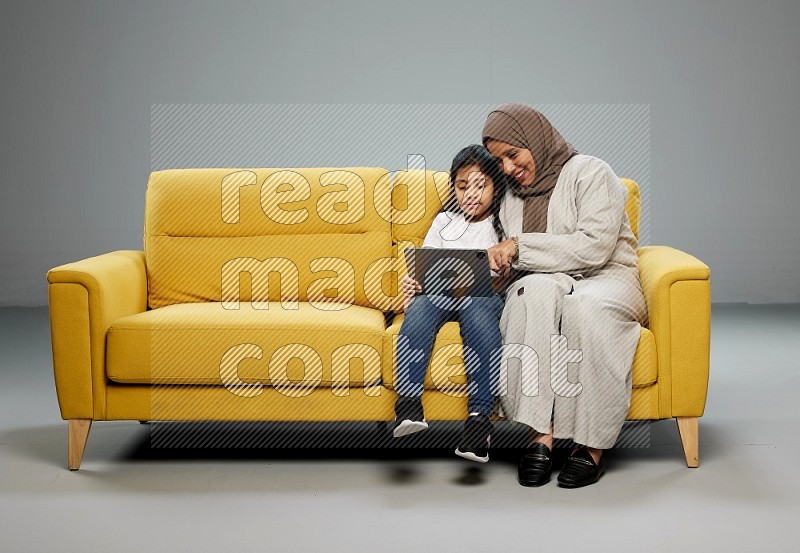 A girl with her mother on yellow sofa and watching on iPad on gray background