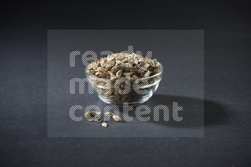 A glass bowl full of cardamom and more seeds spreaded beneath the bowl on black flooring