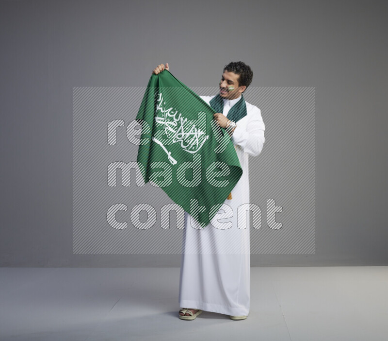 A Saudi man standing wearing thob with face painting wrapping Saudi flag scarf and holding big Saudi flag on gray background