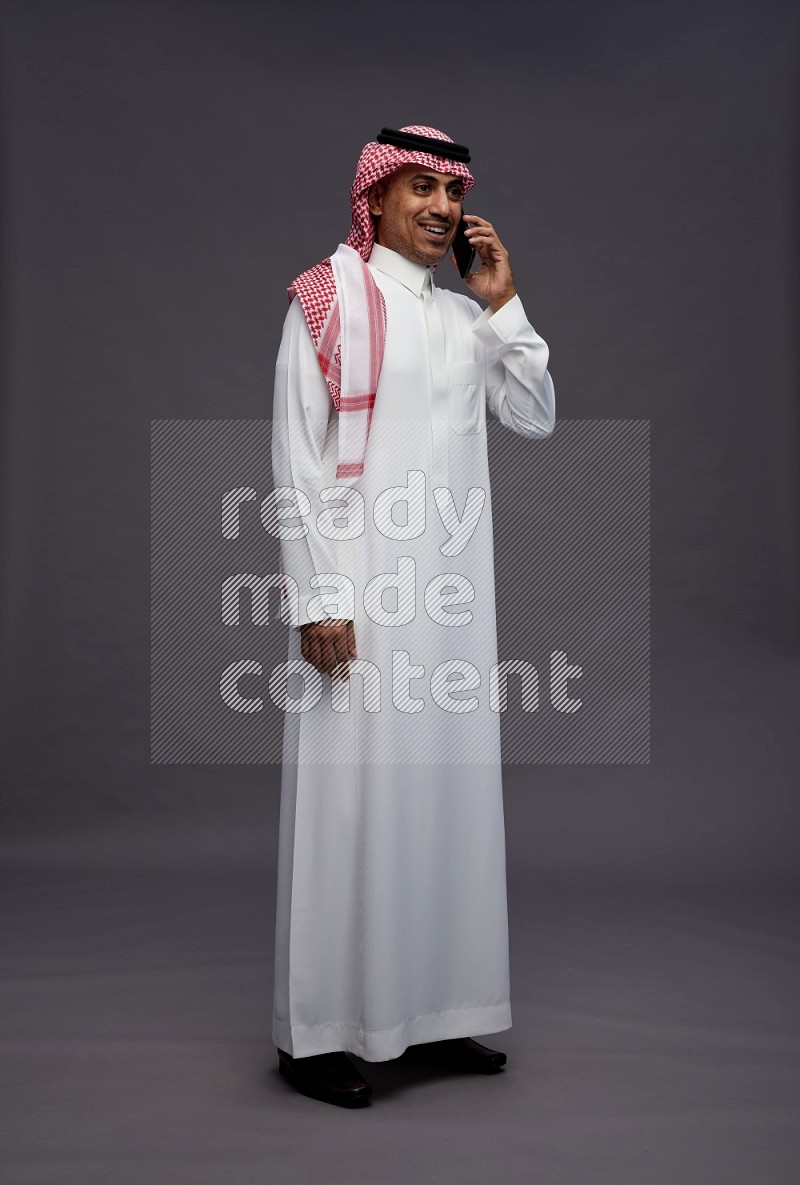 Saudi man wearing thob and shomag standing talking on phone on gray background