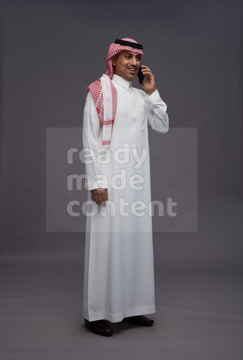 Saudi man wearing thob and shomag standing talking on phone on gray background