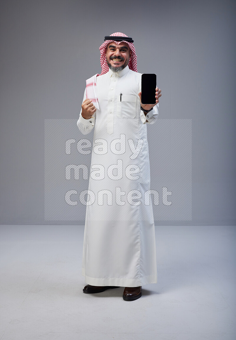 Saudi man Wearing Thob and red Shomag standing showing phone to camera on Gray background