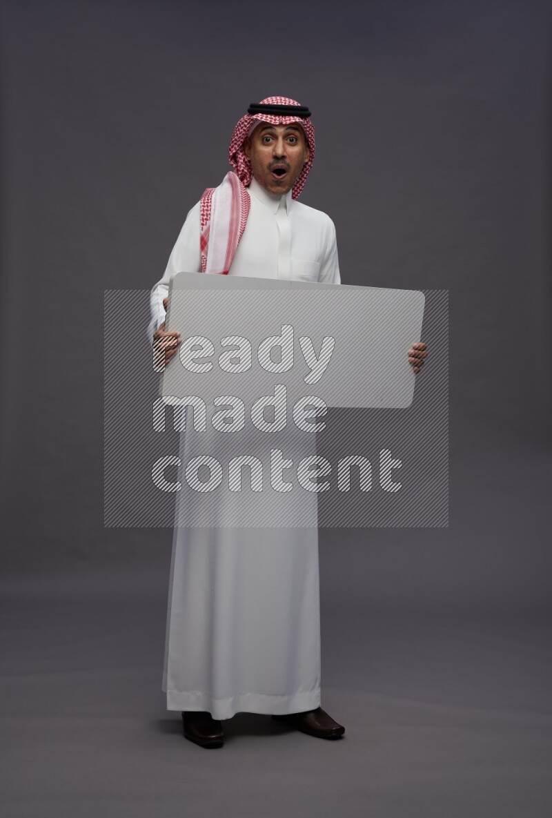 Saudi man wearing thob and shomag standing holding board on gray background