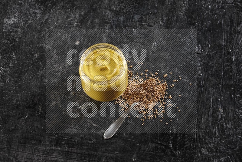 A glass jar full of mustard paste and a metal spoon full of mustard seeds on a textured black flooring