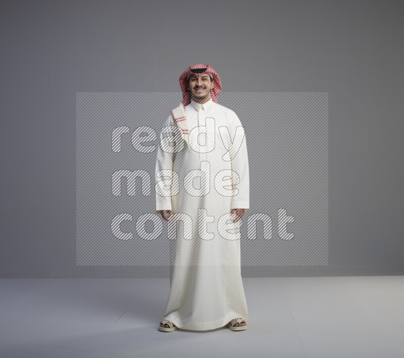 A Saudi man standing wearing thob and red shomag interacting with the camera on gray background