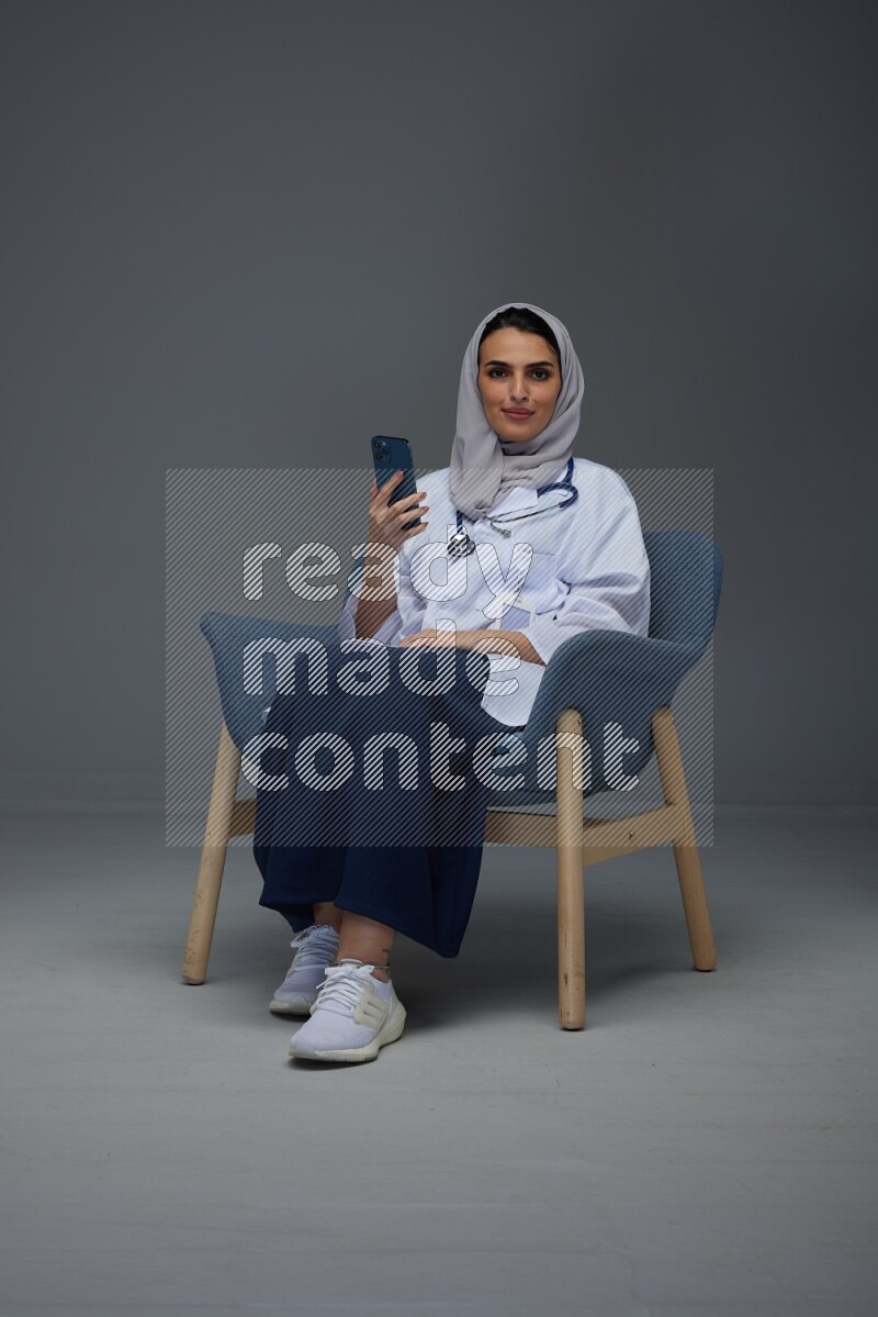 A doctor wearing a light gray head scarf sitting on a dark grey chair and using a laptop while talking in the phone eye level on a grey background