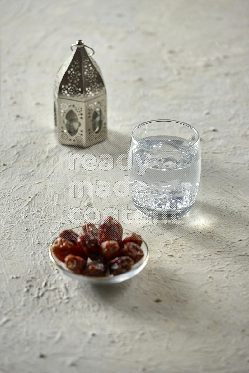 A silver lantern with drinks, dates, nuts, prayer beads and quran on textured white background