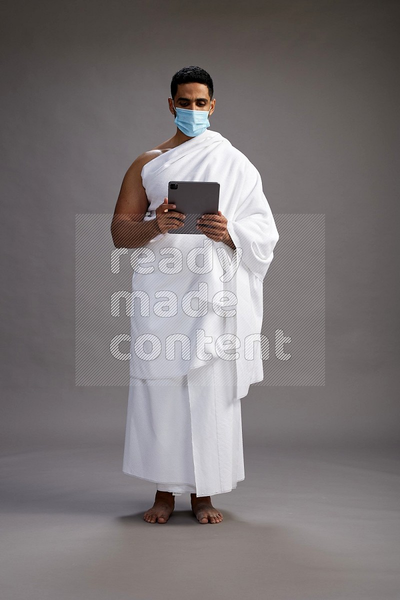 A man wearing Ehram with face mask Standing holding a tablet on gray background