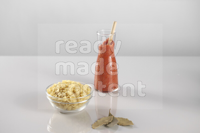 Raw pasta with tomatoe pasta with different ingredients such as cherry tomatoes, basil, garlic, bay laurel, cardamom, white pepper, black pepper, red chilis and wheat stalks on light grey background