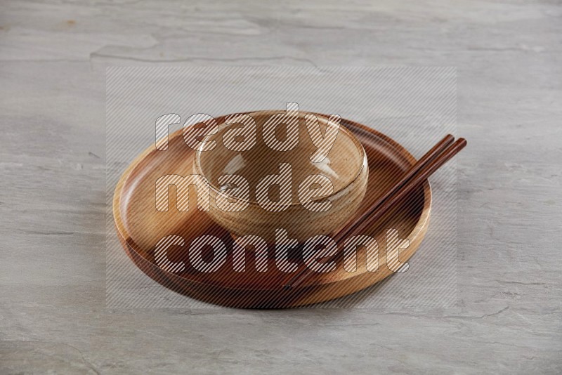 multi color pottery round bowl on top of brown wood round plate and wood chopsticks, on grey textured countertop