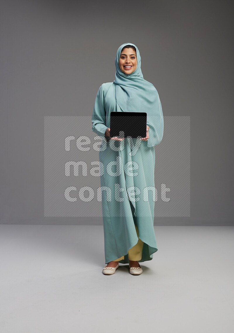 Saudi Woman wearing Abaya standing showing tablet to camera on Gray background