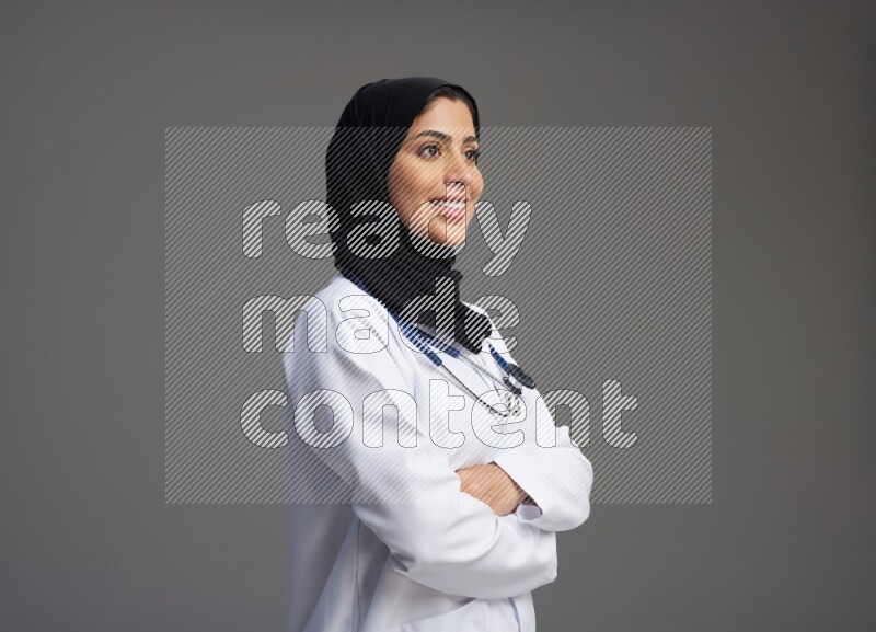 Saudi woman wearing lab coat with stethoscope standing with crossed arms on Gray background