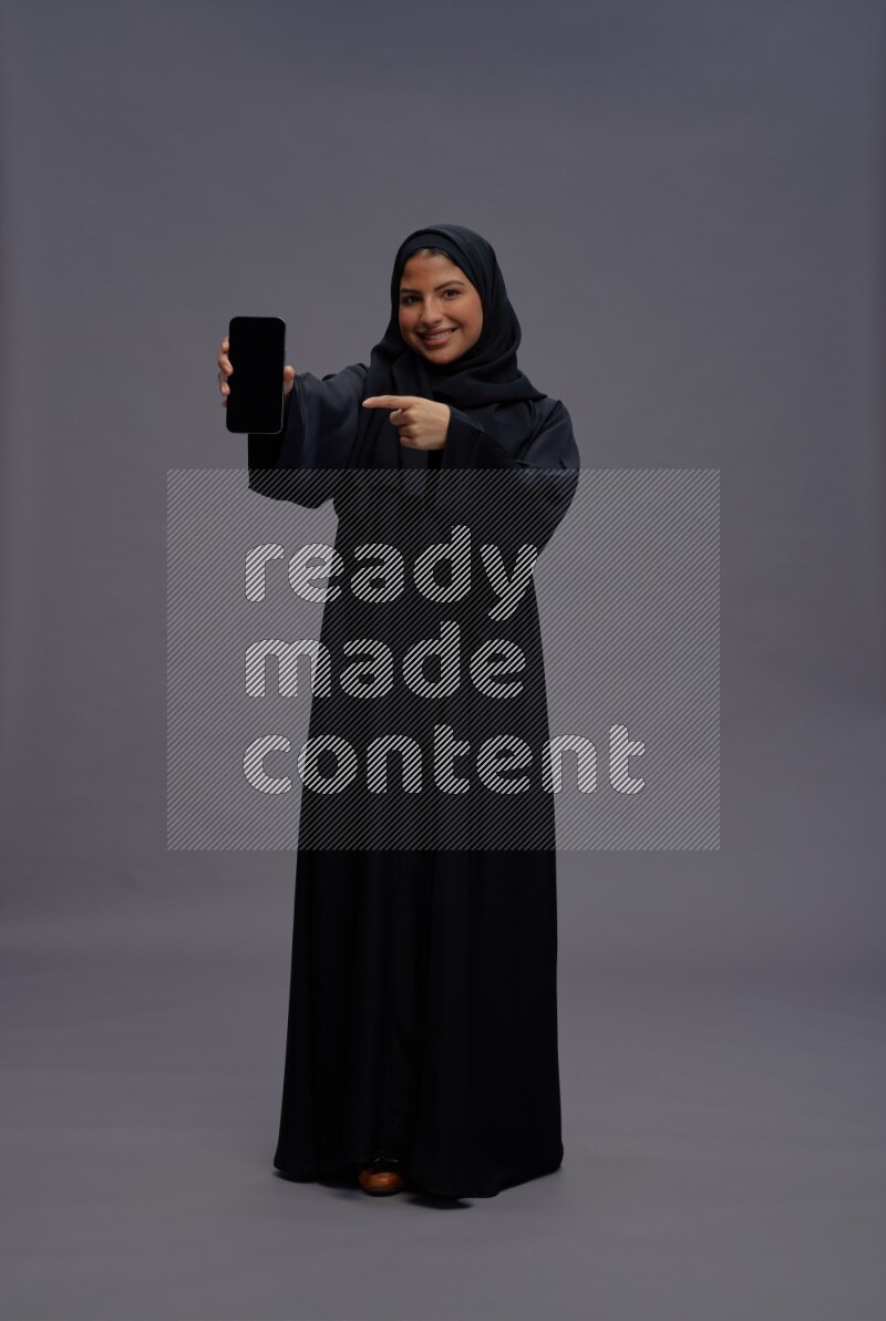 Saudi woman wearing Abaya standing showing phone to camera on gray background