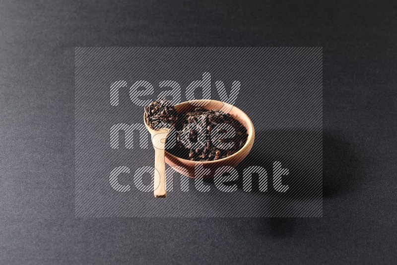 A wooden bowl and a wooden spoon full of cloves on a black flooring
