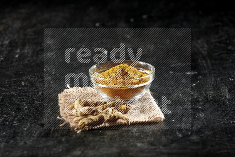 A glass bowl full of turmeric powder with dried turmeric fingers on a burlap fabric on textured black flooring