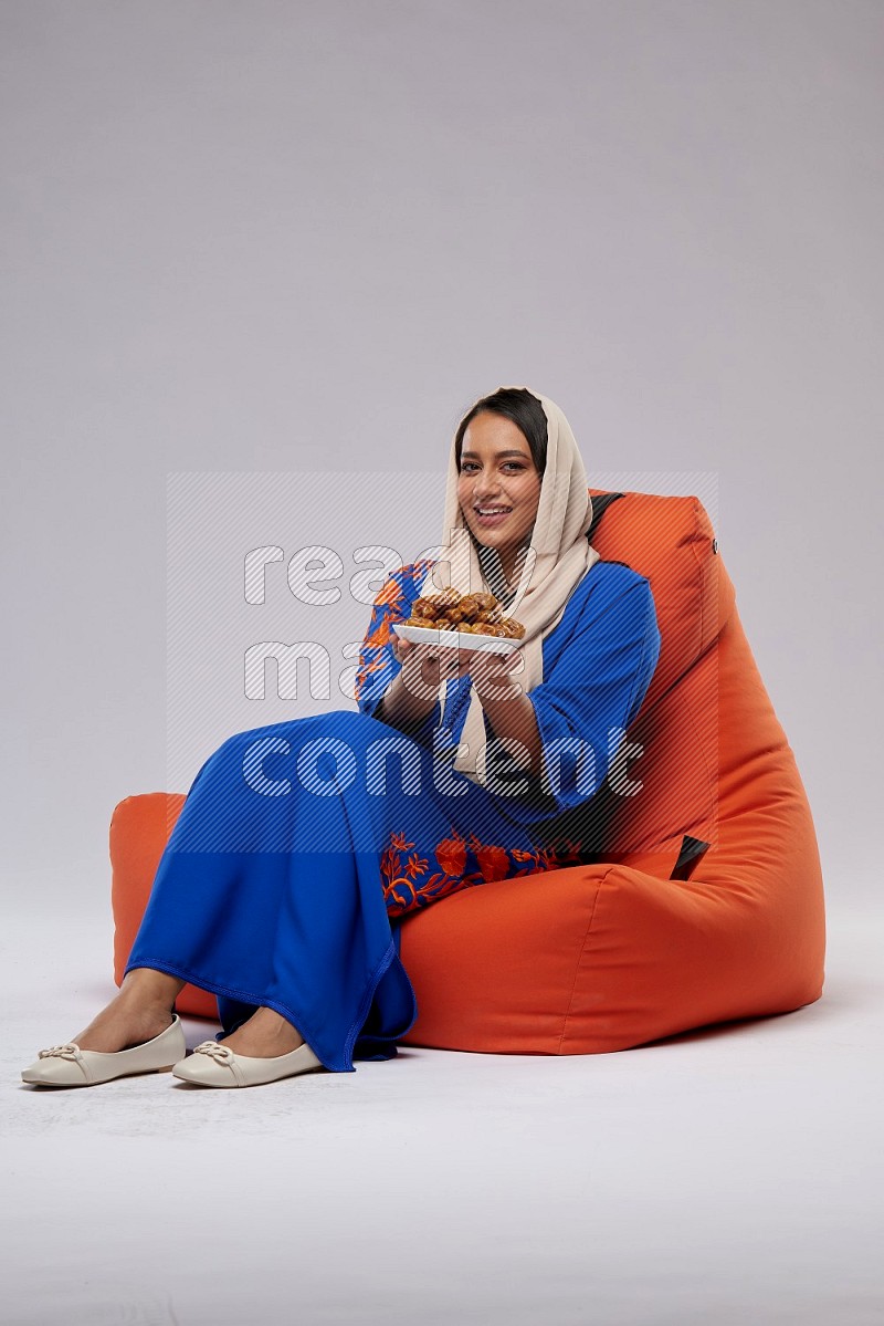 A Woman sitting on an orange beanbag wearing Jalabeya holding a plate of dates