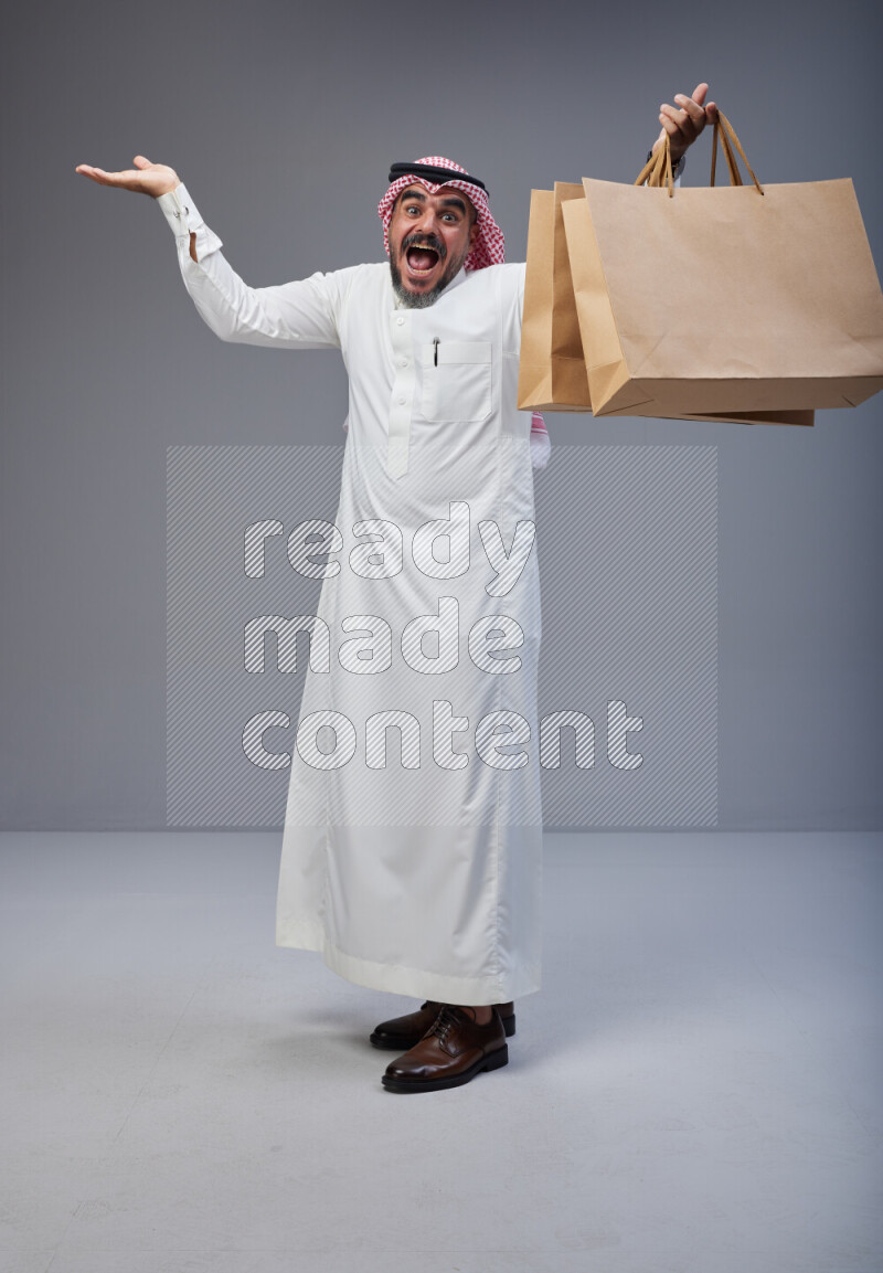 Saudi man Wearing Thob and red Shomag standing holding shopping bag on Gray background