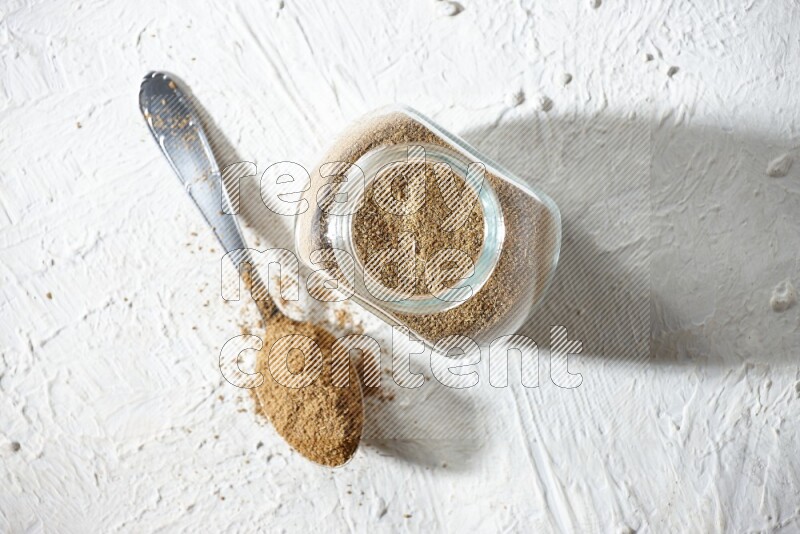 وعاء زجاجي للتوابل وملعقة معدنية ممتلئان ببودرة الكمون على خلفية بيضاء