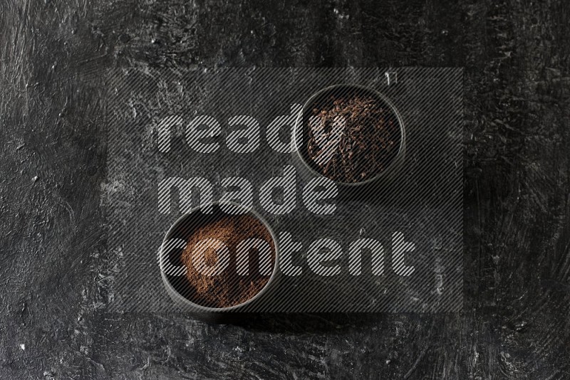 2 Black pottery bowls full of cloves and the other full of cloves powder on textured black flooring