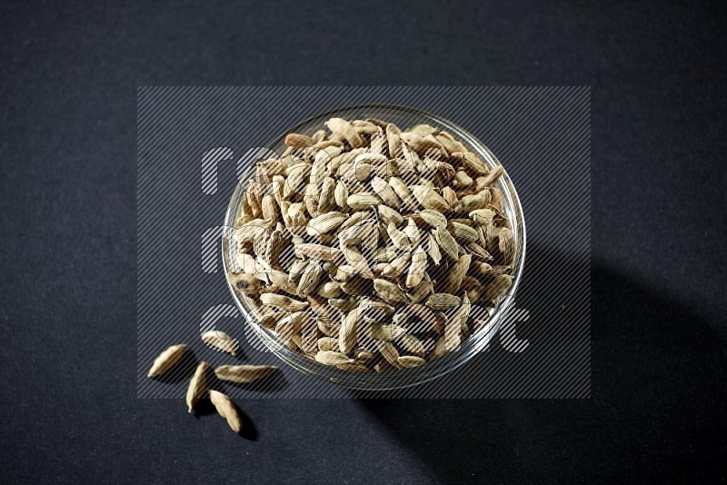 A glass bowl full of cardamom and more seeds spreaded beneath the bowl on black flooring