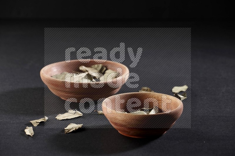 2 wooden bowls full of dried bay leaves with more leaves spread on black flooring